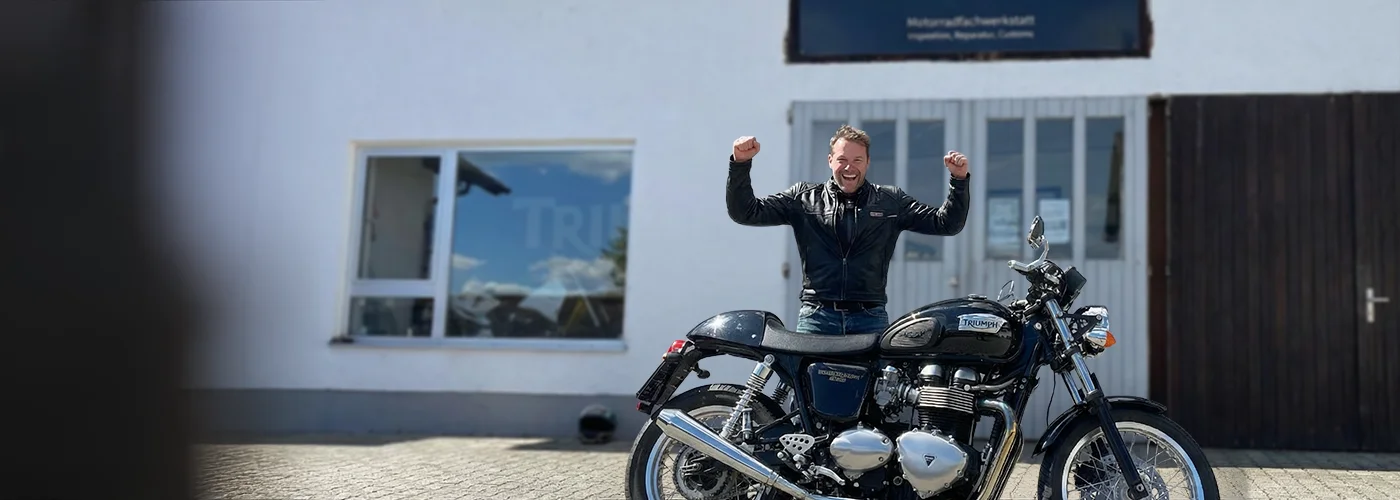 Glücklicher Kunde mit erhobenen Armen neben einer Triumph Thruxton Café Racer vor unserer Motorradwerkstatt in Emmering bei München.